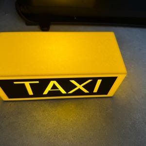 May include: A rectangular yellow taxi roof light with the word "TAXI" in bold, yellow letters on a black background. The light is illuminated, suggesting it is turned on.