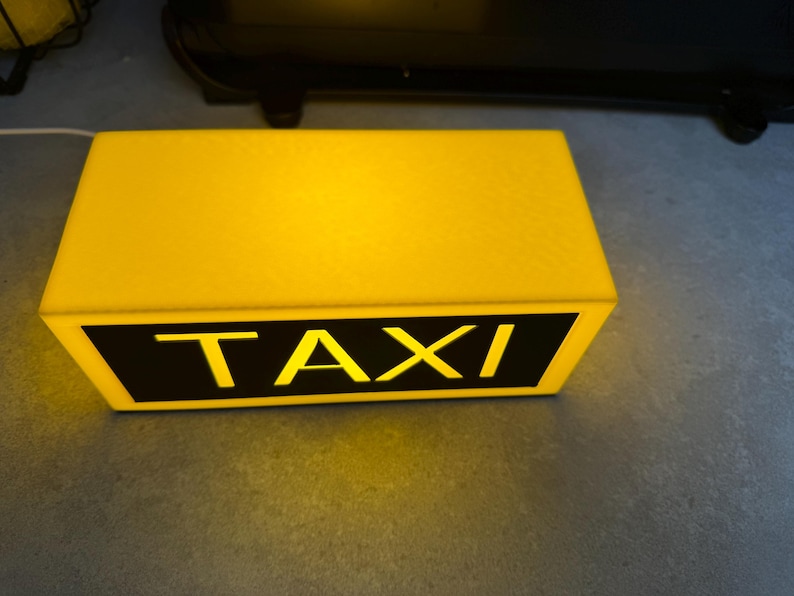 May include: A rectangular yellow taxi roof light with the word "TAXI" in bold, yellow letters on a black background. The light is illuminated, suggesting it is turned on.