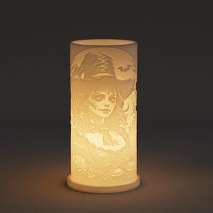 May include: A cylindrical, off-white ceramic lamp with a carved design featuring a woman in a witch hat, bats, and pumpkins. The lamp is illuminated from within, casting a warm glow. The design is detailed and intricate, perfect for Halloween.