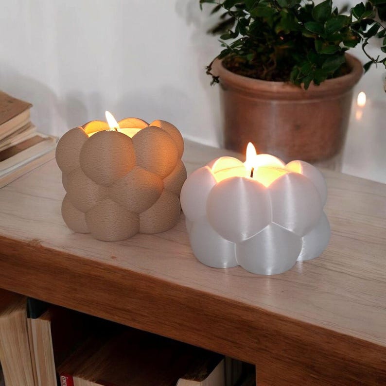 May include: Two bubble-shaped candles with lit flames. One candle is beige, the other is white. The candles sit on a wooden surface. A potted plant is in the background.