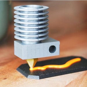 May include: A silver 3D printer hot end with a series of stacked cooling fins. The hot end is dispensing a bright orange filament onto a black, hexagonal surface, creating a wavy line. The setup is on a wooden surface.