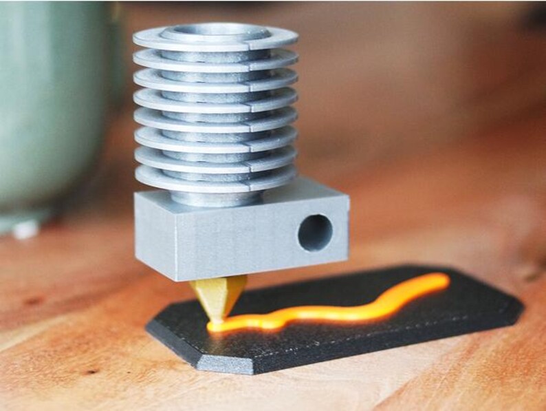 May include: A silver 3D printer hot end with a series of stacked cooling fins. The hot end is dispensing a bright orange filament onto a black, hexagonal surface, creating a wavy line. The setup is on a wooden surface.