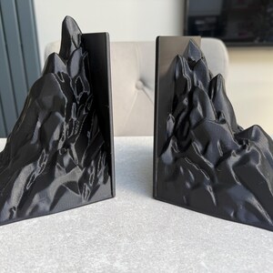 May include: A pair of black mountain-shaped bookends. Each bookend features a detailed, textured design resembling a rugged mountain range. The bookends are made of a solid, dark material and are designed to hold books upright.