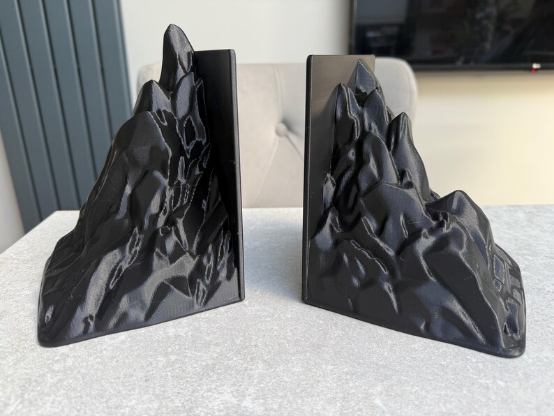 May include: A pair of black mountain-shaped bookends. Each bookend features a detailed, textured design resembling a rugged mountain range. The bookends are made of a solid, dark material and are designed to hold books upright.