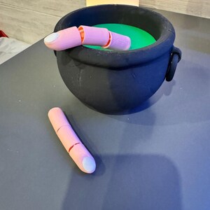 May include: A black cauldron filled with green liquid, with a pink finger prop partially submerged. Another detached pink finger prop lies nearby. The props have a realistic design, perfect for Halloween or themed events.