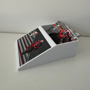May include: White desk organiser with a slanted top and a rectangular base. The organiser features a racing theme with images of red motorcycles on a track. The text "moto rapido" is visible on the images.