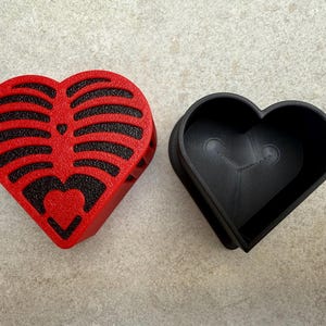 May include: A red heart-shaped box with a black rib cage design on the lid, next to a black heart-shaped container. The box is open, revealing the interior. The design is a unique and artistic representation.