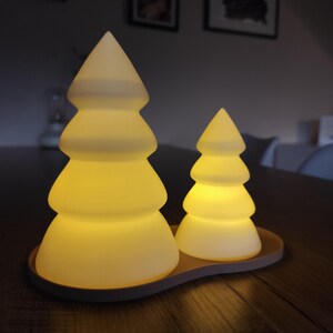 May include: Two white ceramic Christmas tree night lights with a warm yellow glow, sitting on a brown base.