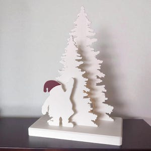 May include: A white wooden Christmas decoration featuring a stylized Santa Claus figure with a burgundy hat, waving at a layered white Christmas tree. The decoration is mounted on a white rectangular base.