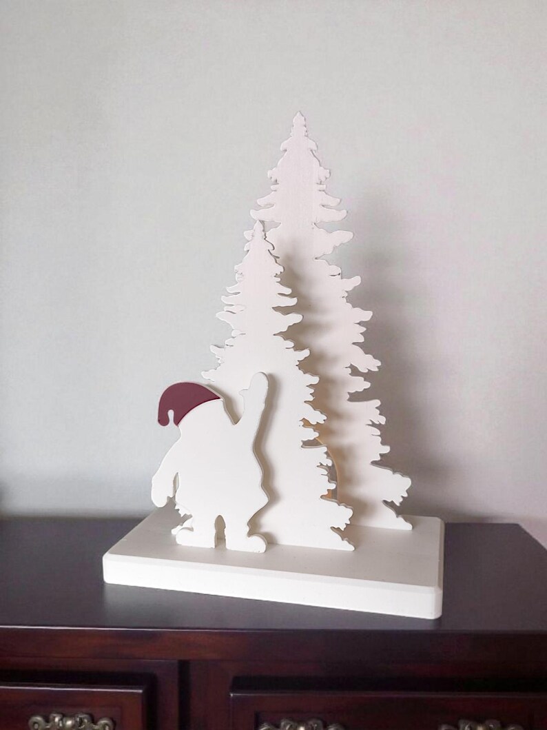 May include: A white wooden Christmas decoration featuring a stylized Santa Claus figure with a burgundy hat, waving at a layered white Christmas tree. The decoration is mounted on a white rectangular base.