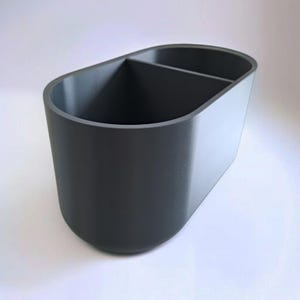 Puede incluir: Un organizador de escritorio gris oscuro, de forma ovalada, con dos compartimentos. El organizador est&aacute; hecho de un material s&oacute;lido y tiene un acabado liso. El dise&ntilde;o es simple y moderno, adecuado para guardar bol&iacute;grafos, l&aacute;pices u otros &uacute;tiles de oficina.