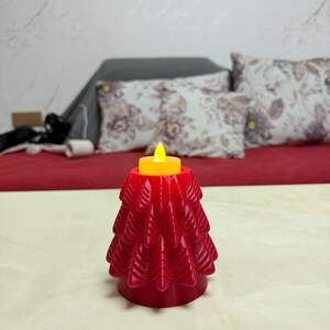 May include: A red, tree-shaped candle holder with a yellow LED candle. The candle holder has a detailed, layered design. The background includes a red surface and decorative pillows.