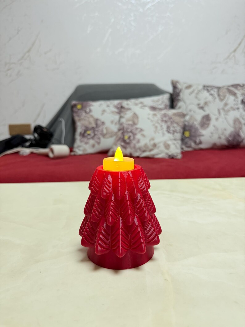 May include: A red, tree-shaped candle holder with a yellow LED candle. The candle holder has a detailed, layered design. The background includes a red surface and decorative pillows.