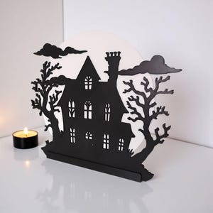 Pu&ograve; includere: Decorazione di Halloween in metallo nero raffigurante la silhouette di una casa infestata con camino, finestre e un albero. La casa &egrave; ambientata su uno sfondo bianco con nuvole nere. Una candela accesa &egrave; in primo piano.