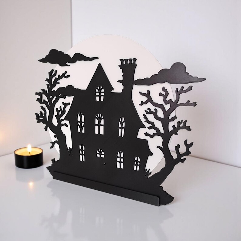 Pu&ograve; includere: Decorazione di Halloween in metallo nero raffigurante la silhouette di una casa infestata con camino, finestre e un albero. La casa &egrave; ambientata su uno sfondo bianco con nuvole nere. Una candela accesa &egrave; in primo piano.