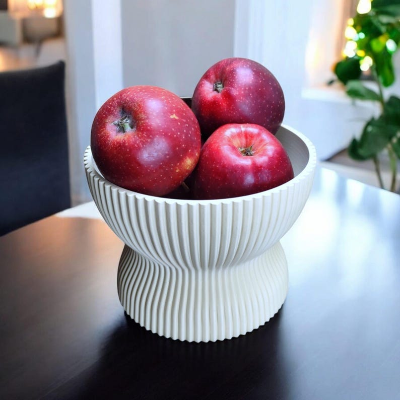 May include: A decorative white bowl with a ribbed design, holding three red apples. The bowl has a pedestal base and sits on a dark surface. The apples are a deep red color.