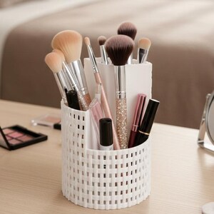May include: A white woven makeup brush holder filled with various brushes, lipsticks, and other cosmetic tools. The brushes have silver and glitter handles. Makeup palettes and a mirror are also visible.