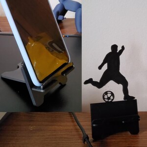 May include: A black phone stand with a phone in it, and a black soccer player silhouette kicking a soccer ball. The phone stand is made of a dark gray material.
