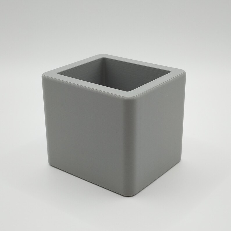 Pu&ograve; includere: Un contenitore quadrato grigio con finitura liscia e opaca. La parte superiore aperta rivela un interno cavo. Gli angoli sono leggermente arrotondati e il design &egrave; minimalista e moderno. Adatto a vari usi.