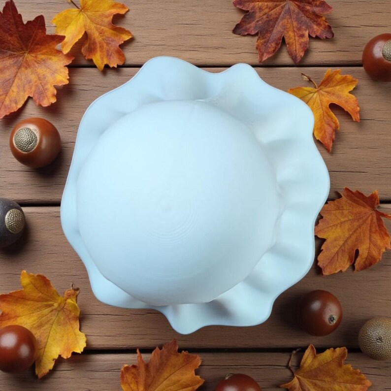 May include: A white, ruffled ceramic bowl with a rounded base. The bowl is surrounded by autumn leaves in shades of orange and brown, and several brown acorns. The arrangement is set on a wooden surface.