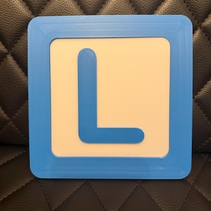 May include: A square sign with a light blue border and a white interior. A large, blue letter "L" is centered on the white background. The sign is likely a learning aid or educational tool.