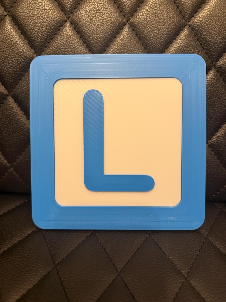 May include: A square sign with a light blue border and a white interior. A large, blue letter "L" is centered on the white background. The sign is likely a learning aid or educational tool.