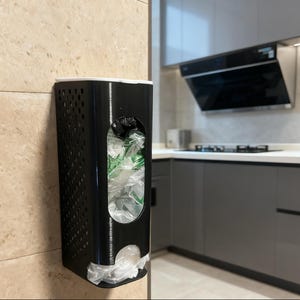 May include: A black plastic bag dispenser mounted on a beige tiled wall. The dispenser has a rectangular shape with a perforated side and a white top. It is filled with plastic bags, and the kitchen is visible in the background.