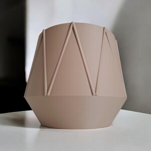 May include: A beige geometric vase with a faceted design. The vase has a cylindrical top and a wider, angular base. Vertical lines create a pattern on the sides. The vase is made of a matte material and sits on a white surface.