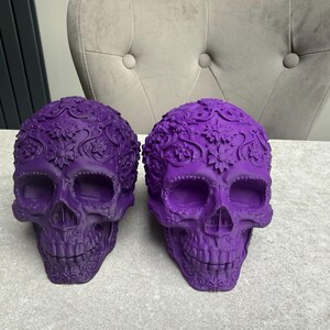 May include: Two decorative purple skull sculptures with floral patterns. The skulls have detailed carvings and are displayed on a light-colored surface. The skulls are a vibrant shade of purple, adding a unique touch to any decor.