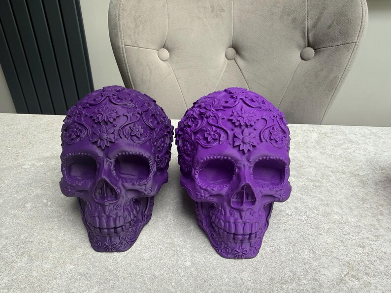 May include: Two decorative purple skull sculptures with floral patterns. The skulls have detailed carvings and are displayed on a light-colored surface. The skulls are a vibrant shade of purple, adding a unique touch to any decor.