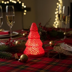 May include: A red, Christmas tree-shaped candle holder with a perforated design sits on a red plaid tablecloth. Champagne flutes and festive decorations surround the tree, creating a holiday atmosphere. The scene is lit with warm, inviting light.