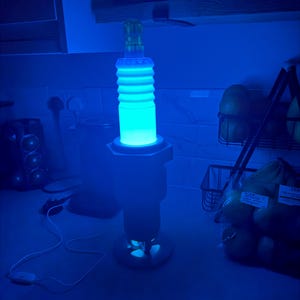 May include: A blue-glowing lamp shaped like a spark plug, with a cylindrical, ribbed body and a base. The lamp emits a bright, cool blue light, creating a futuristic aesthetic. The lamp is plugged into an outlet.