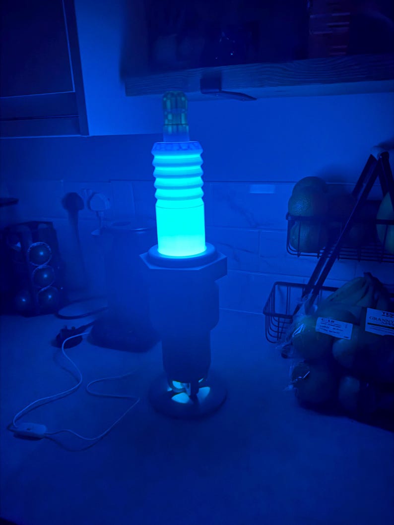 May include: A blue-glowing lamp shaped like a spark plug, with a cylindrical, ribbed body and a base. The lamp emits a bright, cool blue light, creating a futuristic aesthetic. The lamp is plugged into an outlet.