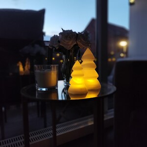 May include: Two white ceramic Christmas tree shaped candle holders with a warm yellow glow. The candle holders are on a black table with a glass of liquid in the background.