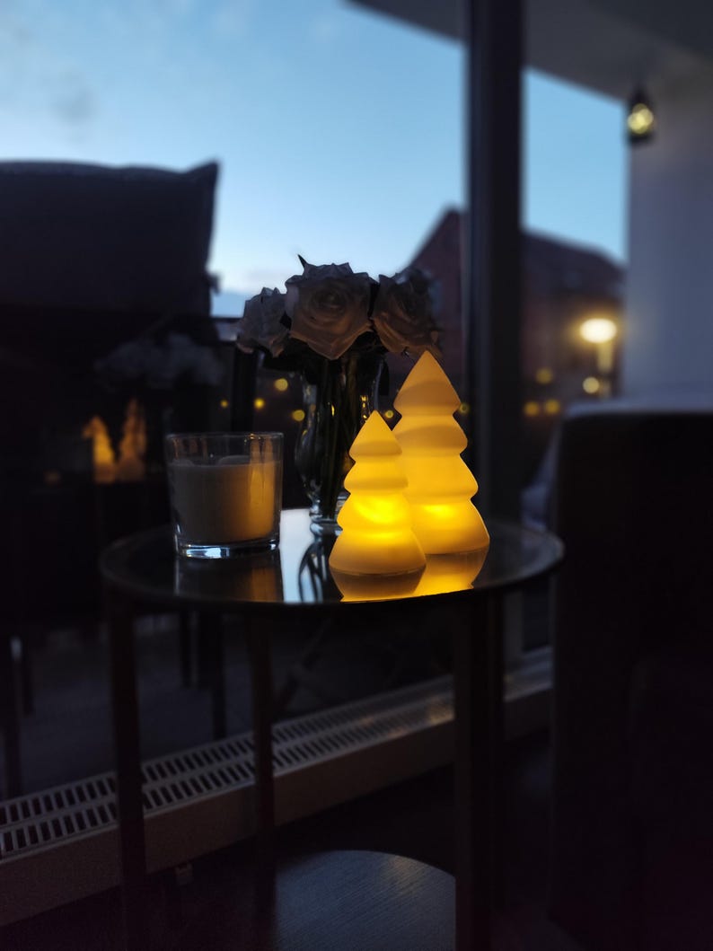 May include: Two white ceramic Christmas tree shaped candle holders with a warm yellow glow. The candle holders are on a black table with a glass of liquid in the background.