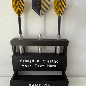 May include: A black dart holder with three darts. The darts have yellow and black striped flights and the text "THE NUKE". The holder has the text "Prim3d & Creat3d Your Text Here" and "GAME ON".