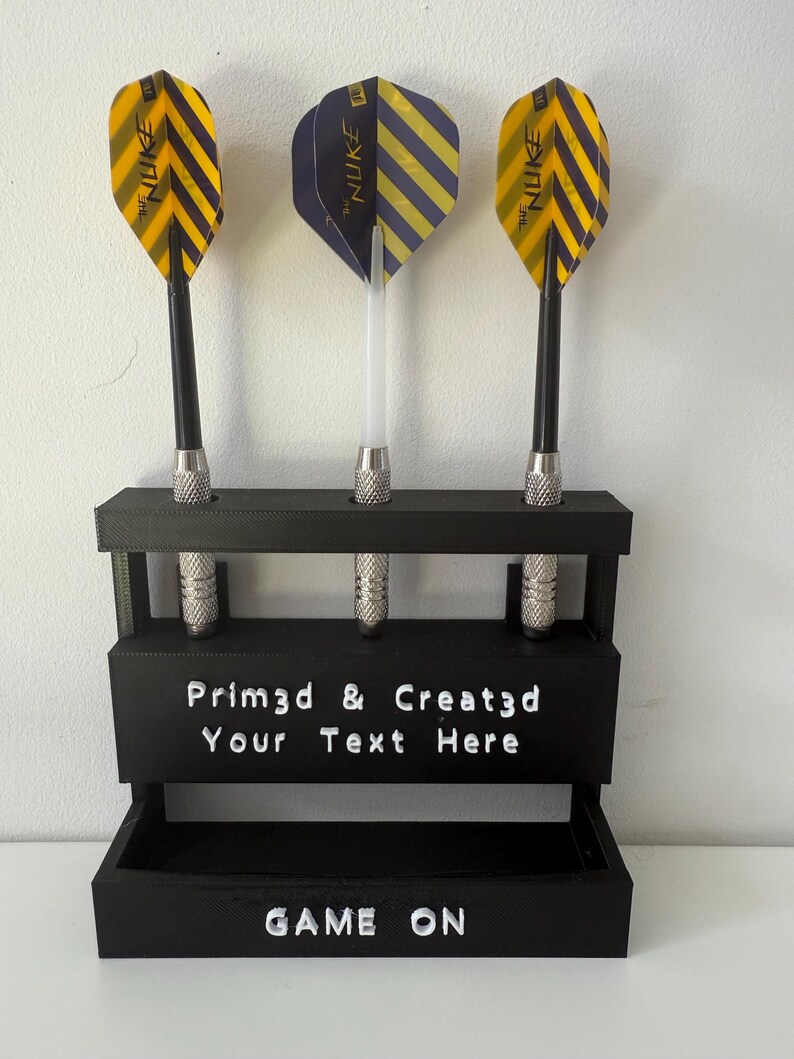 May include: A black dart holder with three darts. The darts have yellow and black striped flights and the text "THE NUKE". The holder has the text "Prim3d & Creat3d Your Text Here" and "GAME ON".