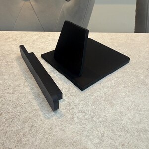 May include: A black wooden phone stand with a rectangular base and a vertical support. A separate black wooden piece is positioned to the left. The stand is designed to hold a phone or tablet. The items are on a light-coloured surface.