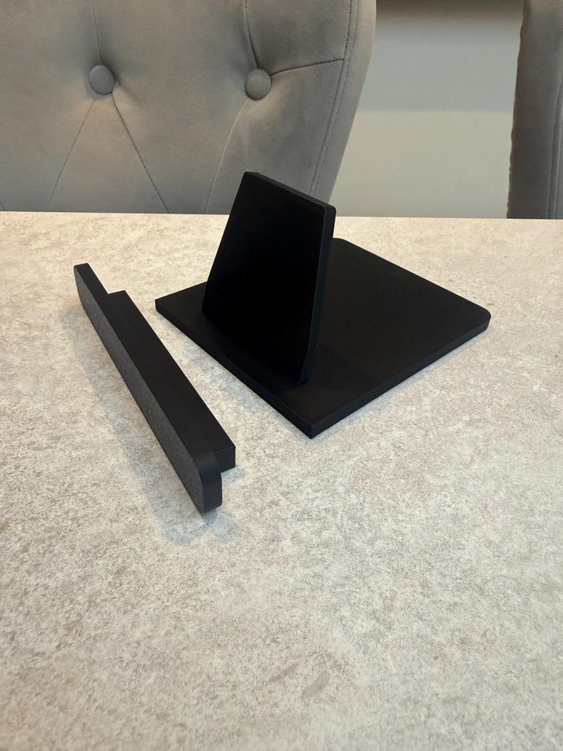 May include: A black wooden phone stand with a rectangular base and a vertical support. A separate black wooden piece is positioned to the left. The stand is designed to hold a phone or tablet. The items are on a light-coloured surface.