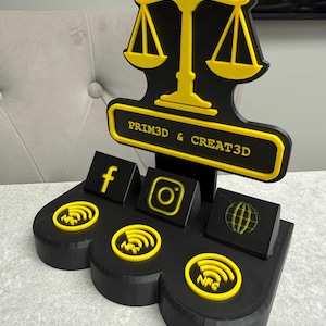 K&ouml;nnte beinhalten: Ein schwarz-gelber 3D-gedruckter Displayst&auml;nder mit einem Waagen-Design. Der St&auml;nder hat Schlitze f&uuml;r Social-Media-Symbole, darunter Facebook, Instagram und einen Globus. Der Text "PRIM3D & CREAT3D" ist auf dem St&auml;nder zu sehen.