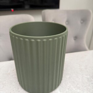 May include: A sage green cylindrical planter with vertical ribbed detailing. The planter is made of a solid material and has a smooth, matte finish. The interior is empty. The background includes a television and two upholstered chairs.