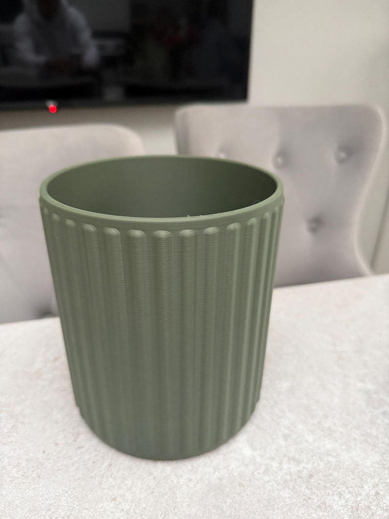 May include: A sage green cylindrical planter with vertical ribbed detailing. The planter is made of a solid material and has a smooth, matte finish. The interior is empty. The background includes a television and two upholstered chairs.