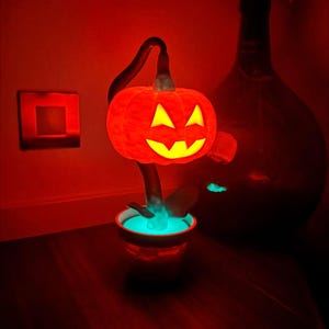 May include: A glowing orange pumpkin-shaped lamp with a carved face, set in a small pot with a turquoise glow. The lamp is on a dark wooden surface, with a red-lit background, creating a Halloween theme.