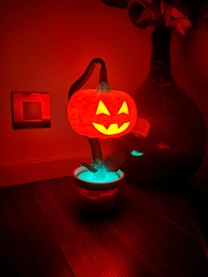 May include: A glowing orange pumpkin-shaped lamp with a carved face, set in a small pot with a turquoise glow. The lamp is on a dark wooden surface, with a red-lit background, creating a Halloween theme.