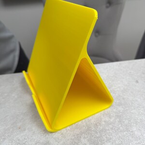 May include: A bright yellow tablet stand with a triangular base and a vertical back. The stand is made of a smooth, solid material and is designed to hold a tablet or phone. The stand is on a light gray surface.