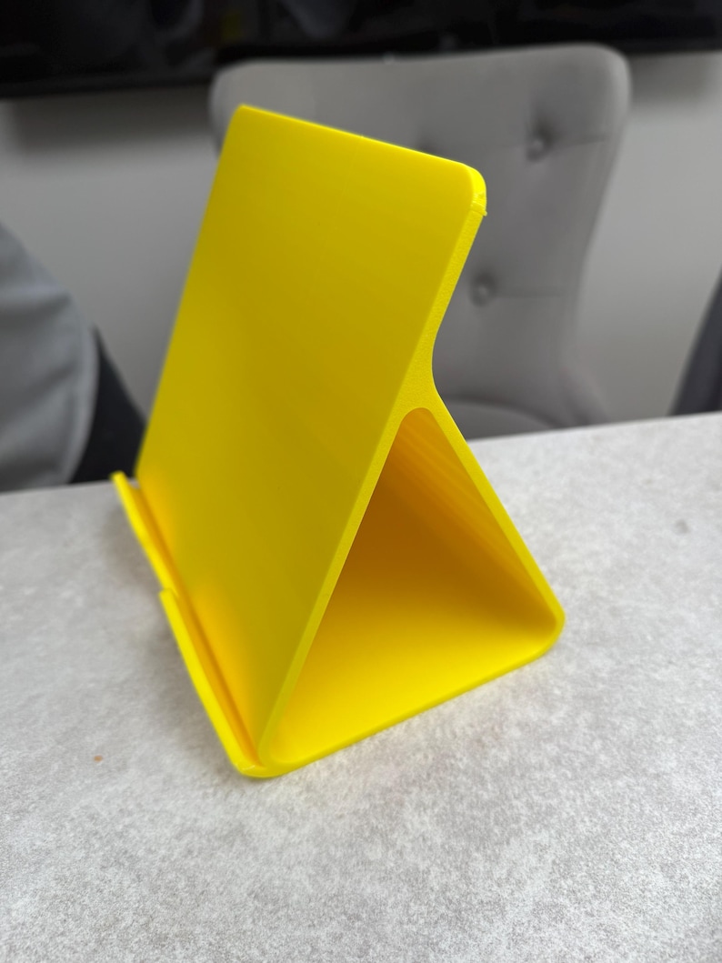 May include: A bright yellow tablet stand with a triangular base and a vertical back. The stand is made of a smooth, solid material and is designed to hold a tablet or phone. The stand is on a light gray surface.