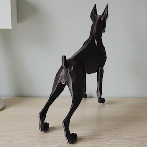 Op de afbeelding: Een zwarte geometrische sculptuur van een hond, waarschijnlijk een Dobermann, op een lichtgekleurd oppervlak. De hond is van achteren te zien, met zijn strakke, hoekige ontwerp en spitse oren. Een modern decoratief stuk.