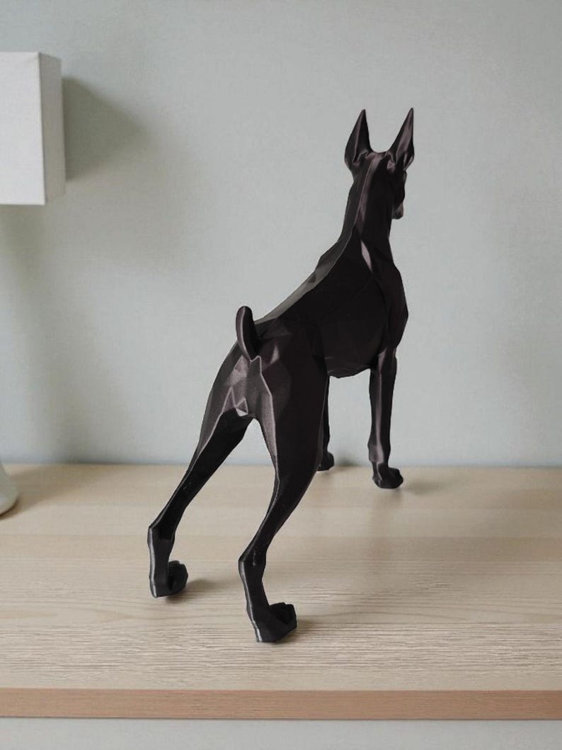 Op de afbeelding: Een zwarte geometrische sculptuur van een hond, waarschijnlijk een Dobermann, op een lichtgekleurd oppervlak. De hond is van achteren te zien, met zijn strakke, hoekige ontwerp en spitse oren. Een modern decoratief stuk.