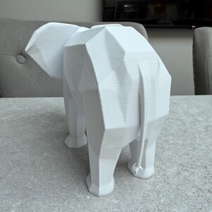Op de afbeelding: Een witte geometrische olifant sculptuur. De sculptuur is gemaakt van hoekige vlakken, waardoor het een moderne, abstracte uitstraling heeft. De olifant staat op een lichtgrijze ondergrond. Het is een decoratief item.