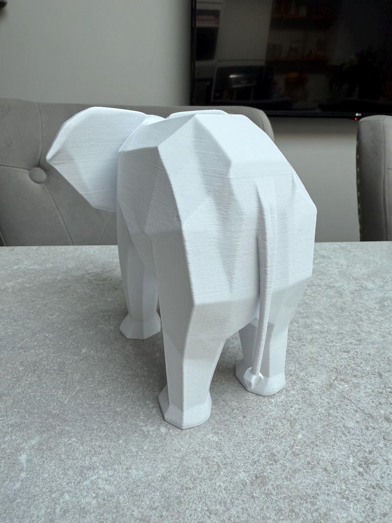 Op de afbeelding: Een witte geometrische olifant sculptuur. De sculptuur is gemaakt van hoekige vlakken, waardoor het een moderne, abstracte uitstraling heeft. De olifant staat op een lichtgrijze ondergrond. Het is een decoratief item.